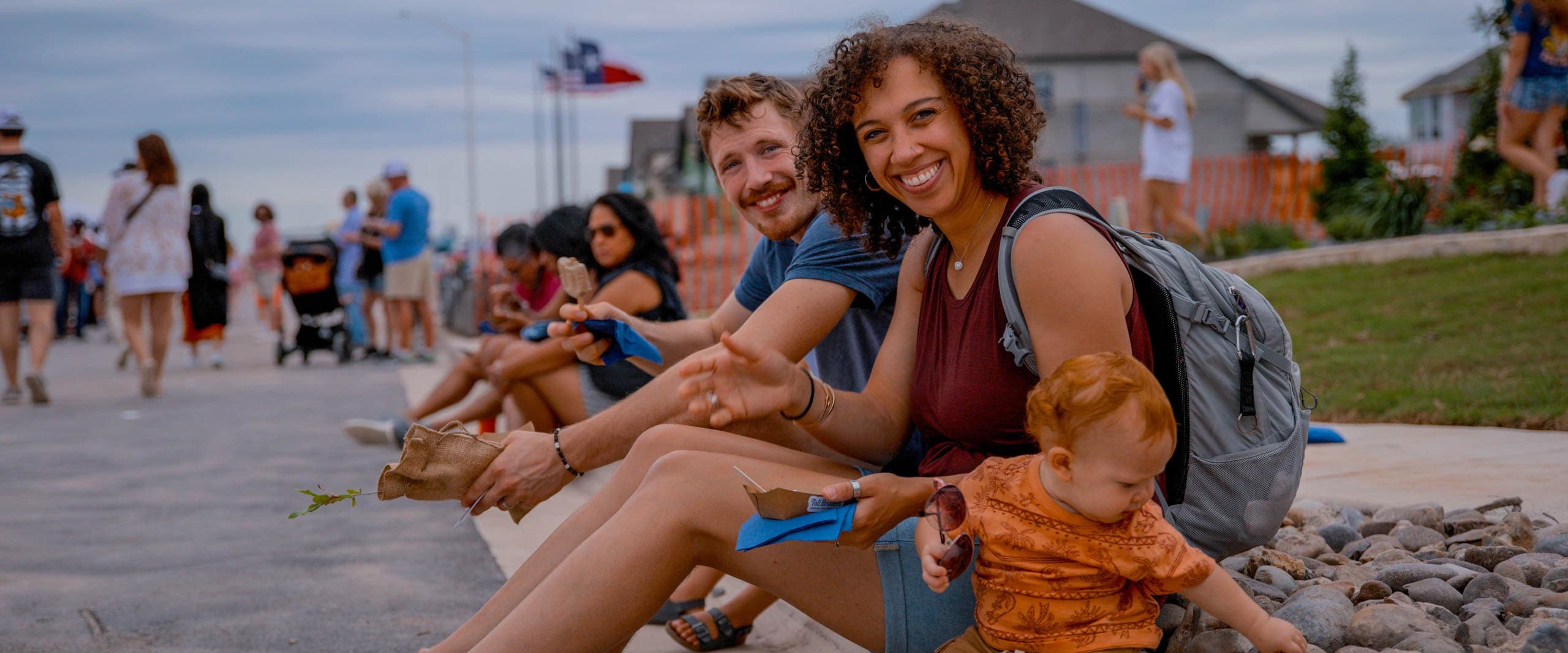 A group of people, including a smiling woman holding a baby, sit on a curb at an outdoor event. Other attendees and flags are visible in the background.