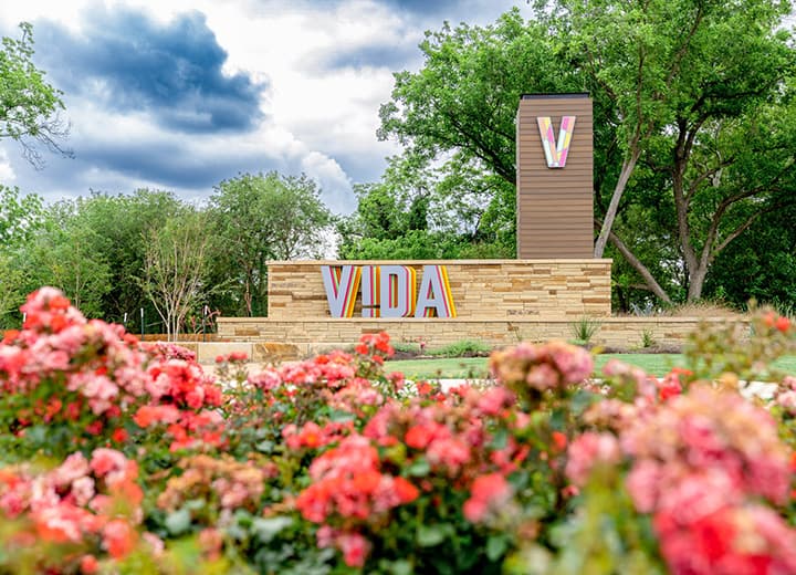 A colorful sign with the word 'vida' set against a backdrop of lush greenery and bright pink flowers in the foreground.