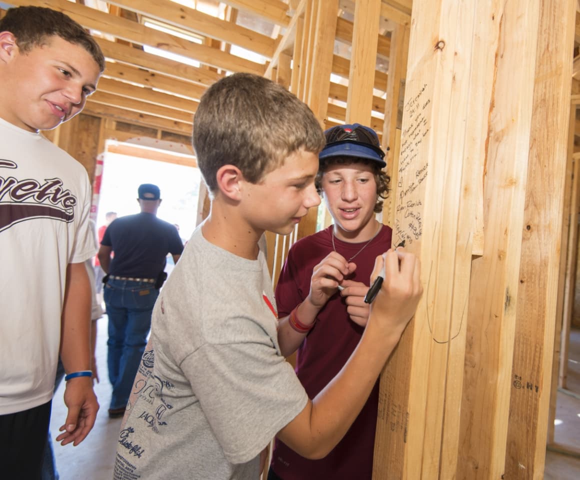 A boy wearing a white t-shirt and signing the wood frame of a home being built.