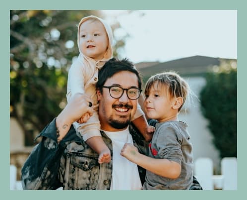 A man with glasses holding two children in front of a house.