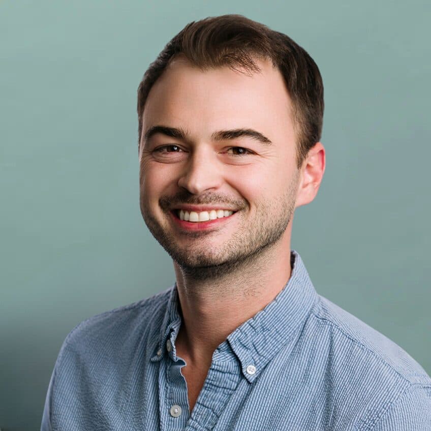 A smiling man with short brown hair and stubble wears a light blue button-up shirt, posing in front of a plain light green background.