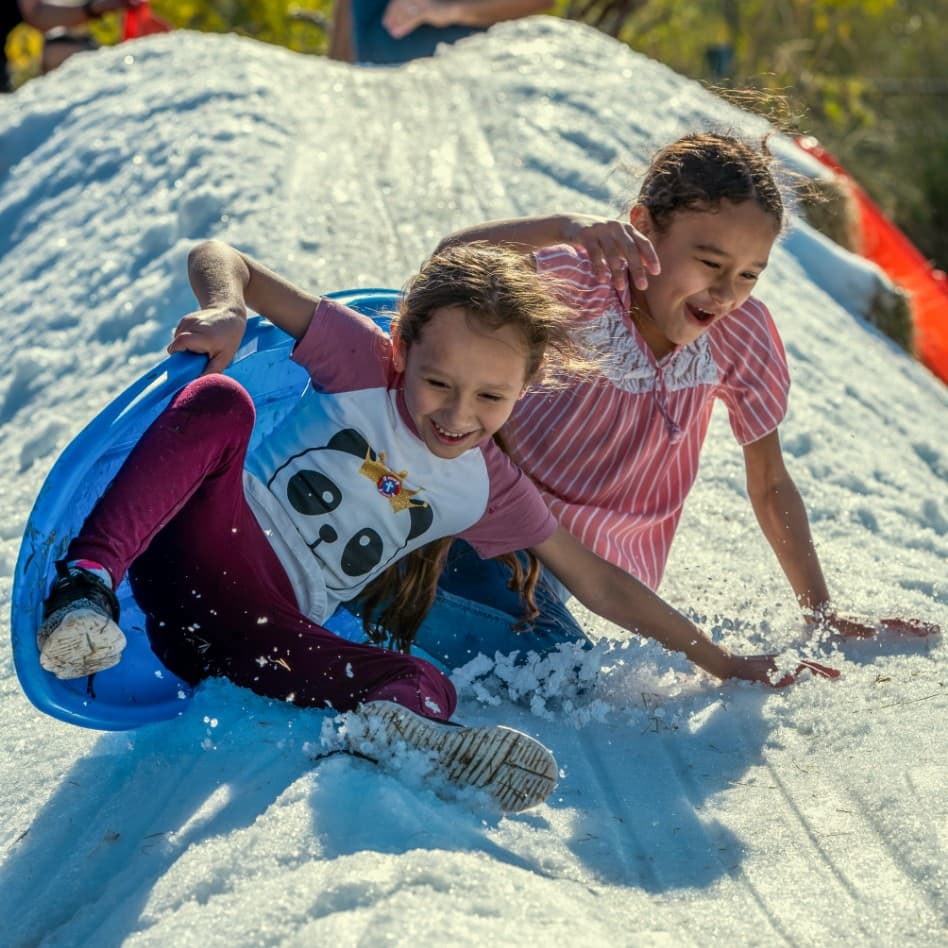 Two girls slide down a snow slide at an outdoor event.