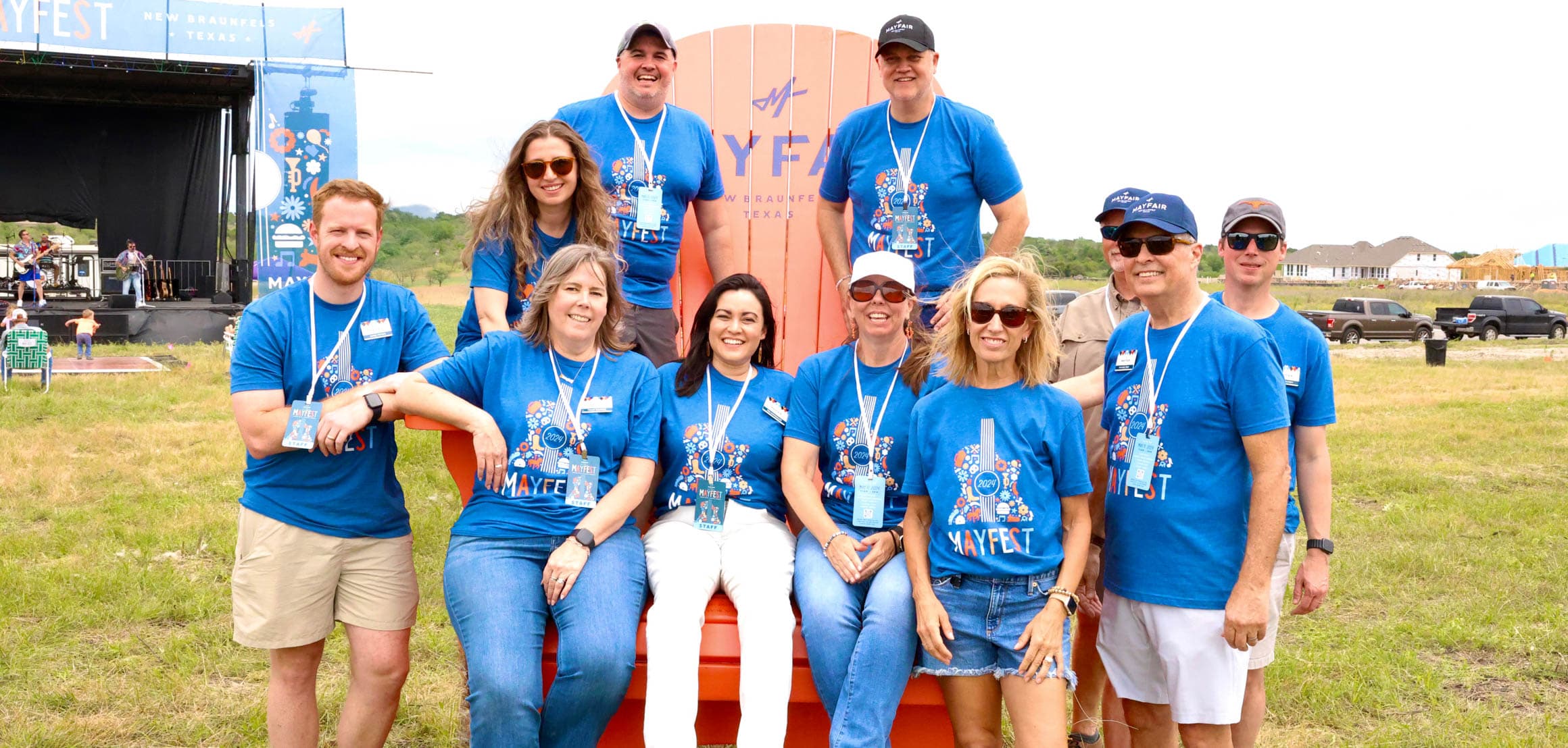 A group of ten people wearing matching blue T-shirts pose outdoors in front of a large orange chair and a stage.