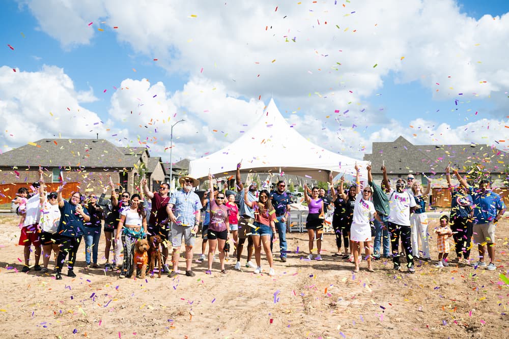 A group of people celebrating outdoors under a white tent with colorful confetti in the air, marking another milestone in VIDA San Antonio's 2025 update. Several houses, symbols of community growth, stand proudly against a blue sky sprinkled with clouds.