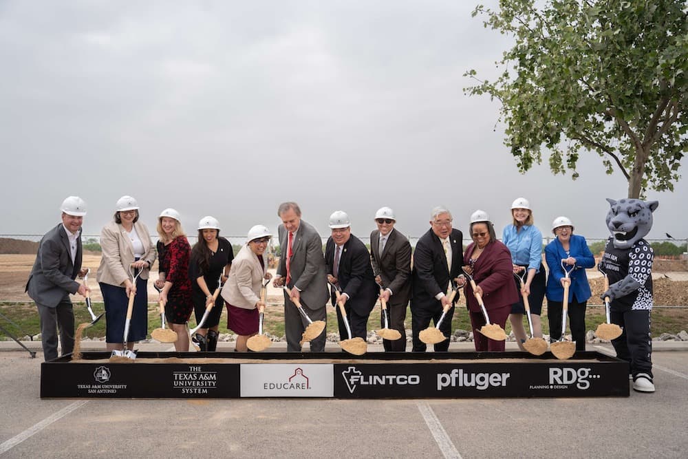 A group of people in hard hats and formal attire, holding shovels and standing in front of a construction site for a groundbreaking ceremony. A mascot stands at the far right.