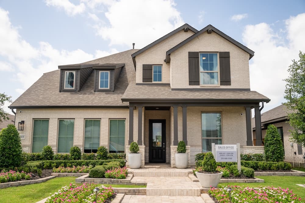 Two-story modern suburban house with brick exterior, black shutters, large windows, manicured lawn, and a sign in front reading "Sales Office.